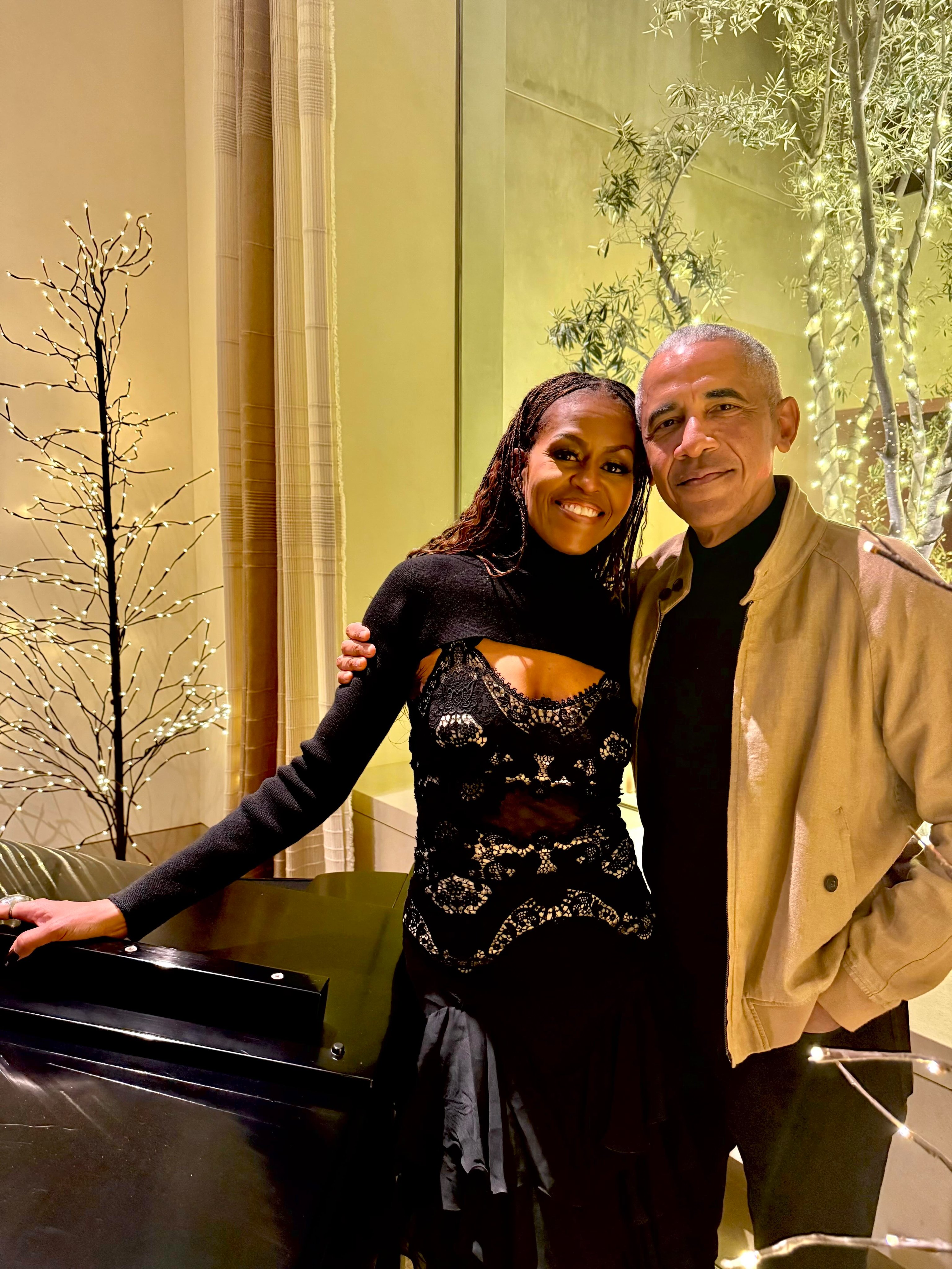 A real image of Barack and Michelle Obama standing near a piano with festive lights behind them. They smile at the camera.